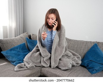 Beautiful Young, Cold Woman Wrapped In A Soft Blanket Sitting On The Couch At Home Talking On The Phone. The Sick Girl Holds A Packet Of Tablets In Her Hand.