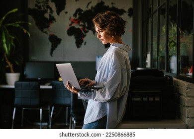 Beautiful young concentrated business woman wearing shirt using laptop while standing in modern workspace - Powered by Shutterstock