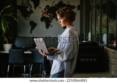 Photo of Beautiful young concentrated business woman wearing shirt using laptop while standing in modern workspace