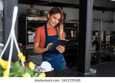 Beautiful young concentrated woman using digital tablet managing a bakery checking online orders in commercial kitchen. - Powered by Shutterstock
