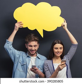 Beautiful young couple in casual clothes is holding a yellow speech bubble, using smartphones and smiling, standing against blackboard