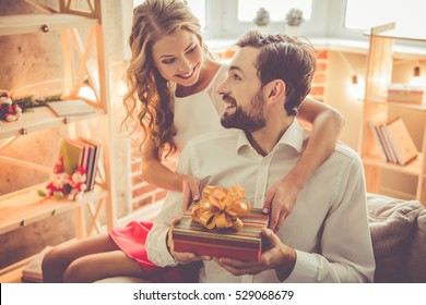 Beautiful Young Couple Is Celebrating At Home. Pretty Girl Is Giving Her Boyfriend A Gift Box