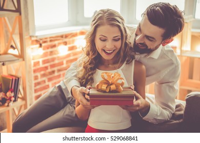 Beautiful young couple is celebrating at home. Handsome man is giving his girlfriend a gift box