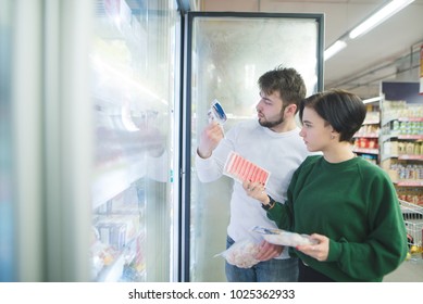 A beautiful young couple chooses frozen foods near the refrigerator in a supermarket. A couple looks at the labels of the goods in the store