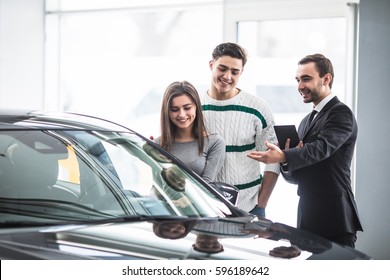Beautiful young couple choosing a car at the dealership talking to the salon manager with tablet in hands