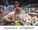 Beautiful young couple is choosing cheese and smiling while doing shopping at the supermarket