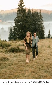 Beautiful Young Couple In Dark Green Clothes Walk By The Hands In The Mountains In Nature. High Quality Photo