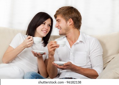 Beautiful Young Couple Drink Coffee Hold Cup, Sitting On A Sofa Happy Smile Looking Each Other Talking, Portrait Of Lovely Young Man And Woman On The Couch