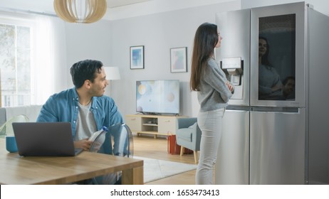 Beautiful Young Couple Drink Morning Coffee in the Kitchen. Girl Checks the Weather Forecast and a To Do List on a Smart Fridge at Home. Apartment is Bright and Cozy.