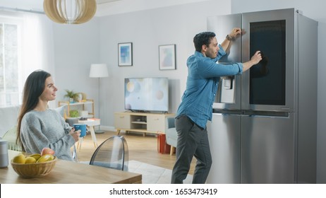 Beautiful Young Couple Drink Morning Coffee in the Kitchen. Male Checks the Weather Forecast and a To Do List on a Smart Fridge at Home. Apartment is Bright and Cozy.