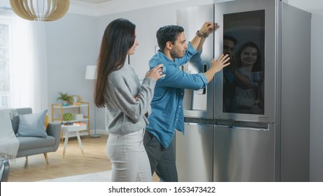 Beautiful Young Couple Drink Morning Coffee in the Kitchen. They Check the Weather Forecast and a To Do List on a Smart Fridge at Home. Apartment is Bright and Cozy.