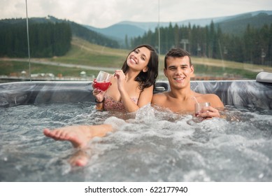 Beautiful young couple enjoying a bath in Jacuzzi while drinking cocktail outdoors on romantic vacation against blurred background of nature