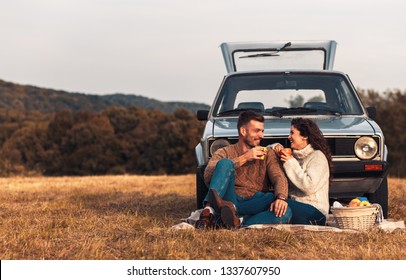 Beautiful young couple enjoying picnic time on the sunset. They drinking tea and sitting in a meadow leaning against a old fashioned car.