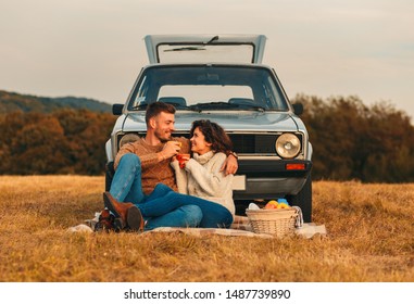 Beautiful young couple enjoying picnic time on the sunset. They drinking tea and sitting in a meadow leaning on old fashioned car.