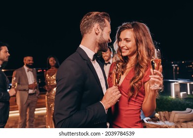 Beautiful young couple in formalwear holding champagne flutes and smiling while spending time on luxury party