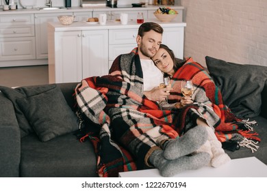 Beautiful Young Couple With Glasses Of White Wine Relaxing On Couch Under Plaid