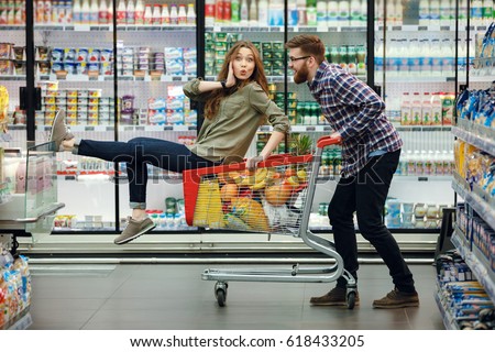 Beautiful young couple having fun while choosing food in the supermarket. Young happy man pushing shopping cart with his girfriend inside