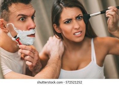 Beautiful Young Couple Having Hectic Morning Habits In Front Of Their Bathroom Mirror. Man Shaving And His Girlfriend Makeup. Selective Focus.