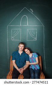 Beautiful Young Couple In House Drawn With Chalk. Affordable Housing For Young Families, Mortgage. Man And Woman Sit On A Sofa Having Embraced. Red-haired Woman And Blonde Man In Check Shirts. 