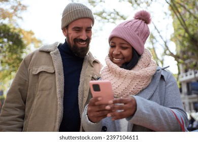 Beautiful young couple interracial in love using mobile phone enjoying winter holiday season in the city streets, walking and hugging and holding gift bags while celebrating Christmas outdoors - Powered by Shutterstock