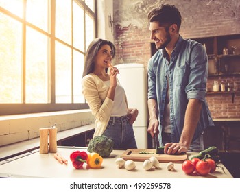 Beautiful Young Couple Is Looking At Each Other And Smiling While Cooking In Kitchen At Home. Man Is Cutting Vegetables