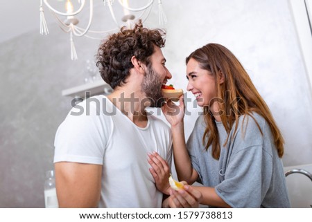Beautiful young couple is looking at each other and feeding each other with smiles while cooking in kitchen at home. Loving joyful young couple embracing and cooking together,having fun in the kitchen