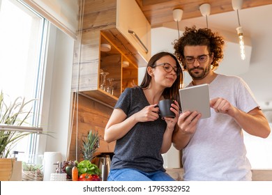 Beautiful young couple in love drinking coffee and reading news using a tablet computer in the kitchen - Powered by Shutterstock