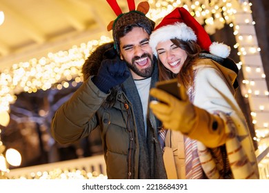 Beautiful young couple in love having video call with friends using smart phone while celebrating New Year in the city streets with Christmas lights all around - Powered by Shutterstock
