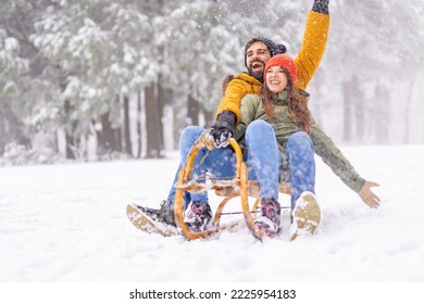 Beautiful young couple in love having fun spending winter vacation in mountains, sitting and hugging on sled, sliding down the hill in the snow