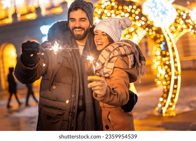 Beautiful young couple in love having fun celebrating New Year on city streets or square, holding sparklers for midnight countdown with Christmas lights in background - Powered by Shutterstock