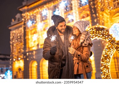 Beautiful young couple in love having fun celebrating New Year on city streets or square, holding sparklers for midnight countdown with Christmas lights in background