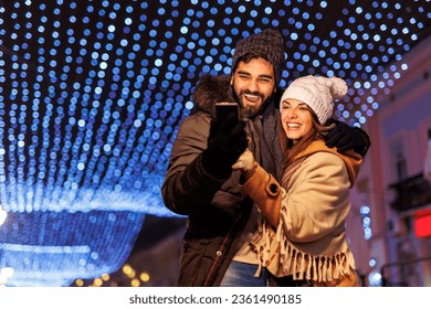 Beautiful young couple in love having video call with friends using smart phone while celebrating New Year's Eve in the city streets with bunch of Christmas lights in the background - Powered by Shutterstock