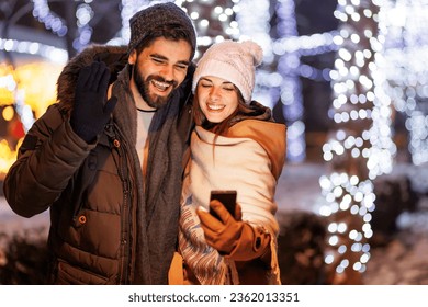 Beautiful young couple in love having video call with friends using smart phone while celebrating New Year's Eve in the city streets with bunch of Christmas lights in the background