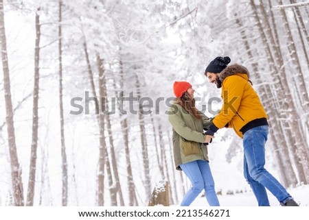 Beautiful young couple in love having fun spending snowy winter day outdoors, holding hands, pulling, pushing and teasing each other while taking a walk through a forest on winter vacation