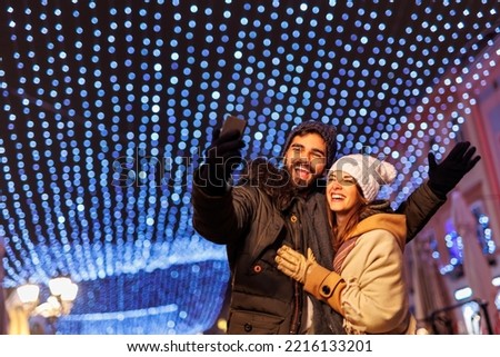 Beautiful young couple in love having fun taking selfies while celebrating New Year's Eve in the city streets with bunch of Christmas lights in the background