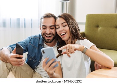 Beautiful young couple in love at home, sitting at the living room, pointing at mobile phone - Powered by Shutterstock