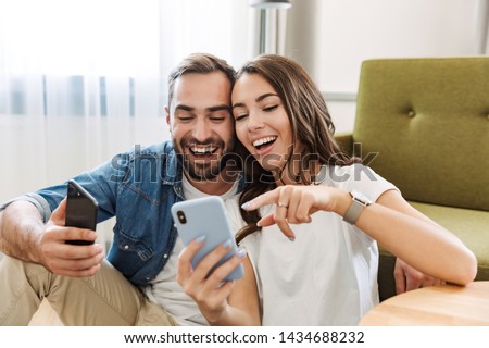 Beautiful young couple in love at home, sitting at the living room, pointing at mobile phone