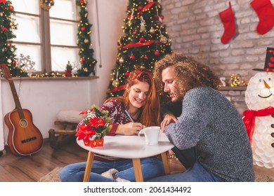 Beautiful young couple in love sitting by the Christmas tree, celebrating Christmas at home, having fun while writing New Years resolutions