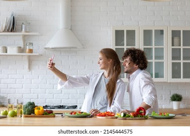 Beautiful Young Couple Making Selfie And Smiling While Preparing Healthy Food In The Kitchen. Capturing Bright Moments Together. 