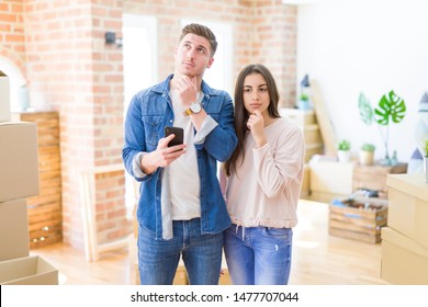 Beautiful young couple moving to a new house using smartphone serious face thinking about question, very confused idea