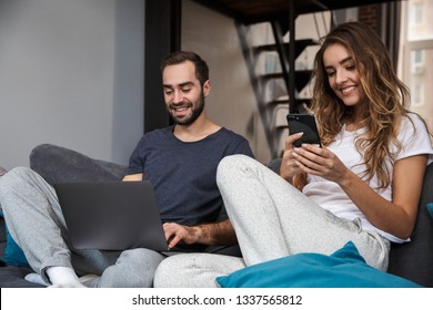 Beautiful Young Couple Relaxing On A Couch At Home, Using Laptop Computer And Mobile Phone