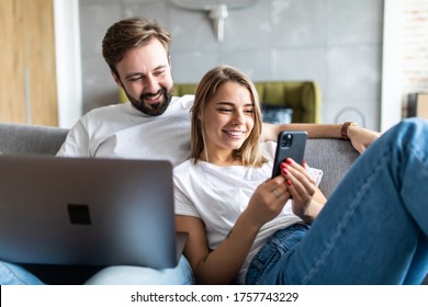 Beautiful Young Couple Relaxing On A Couch At Home, Using Laptop Computer And Mobile Phone