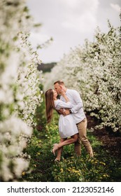 Beautiful Young Couple In A Romantic Place, Spring Blooming Apple Orchard. Happy Joyful Couple Enjoy Each Other While Walking In The Garden. Man Holding Woman's Hand