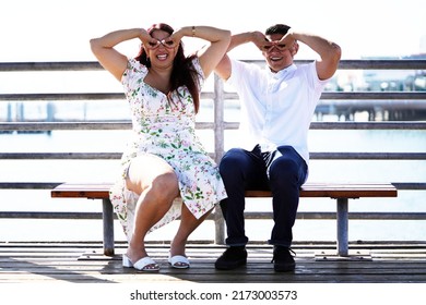 Beautiful young couple sits on a bench overlooking the ocean and makes funny faces while smiling and laughing                              