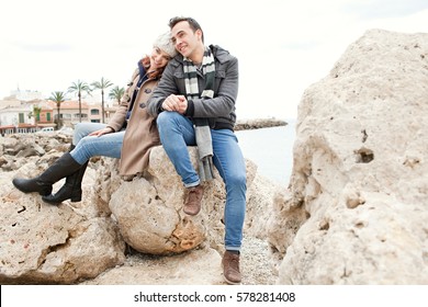 Beautiful young couple sitting on beach rock on winter holiday, wearing coats and hat, smiling joyfully, travel lifestyle outdoors. Boyfriend and girlfriend enjoying seasonal break together, exterior.