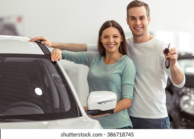 Beautiful young couple is smiling and looking at camera while leaning on their new car in a motor show. Man is holding car keys