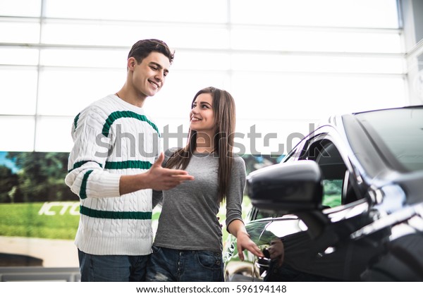 Beautiful young couple standing at\
the dealership choosing the car to buy. Man pointed on\
car.