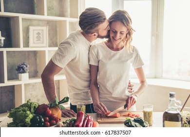 Beautiful young couple is talking and smiling while cooking healthy food in kitchen at home. Man is kissing his girlfriend in cheek - Powered by Shutterstock