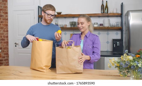 Beautiful Young Couple Unpacking Shopping Bags Full Of Fresh Vegetables And Smiling While Standing In Kitchen Together. Happy Man And Woman Receiving Delivery Groceries At Home