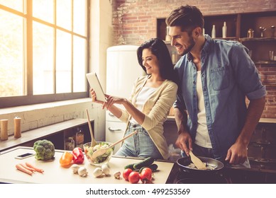 Beautiful young couple is using a digital tablet and smiling while cooking in kitchen at home
